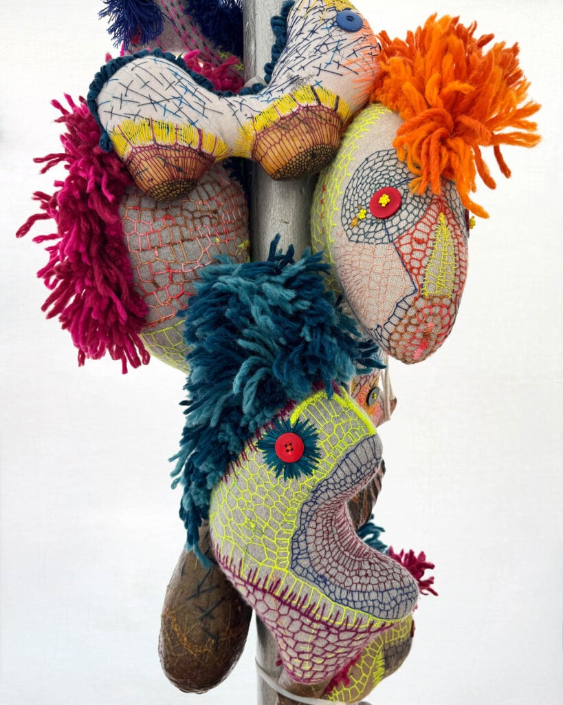 Three colorful, abstract fabric sculptures by Wolfgang Woerner, with textured stitching and yarn hair, are attached to a vertical pole. Each features button eyes and bold patterns.