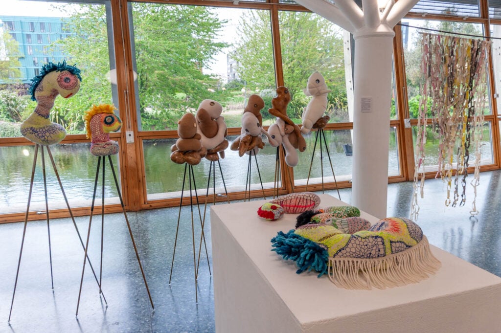 A gallery space displays soft sculptural figures by Wolfgang Woerner on metal stands, with plush, colorful artworks on a white pedestal and beaded strings hanging near large windows overlooking a pond.
