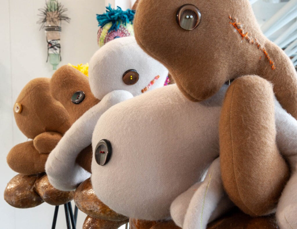 Three large handmade plush creatures by Wolfgang Woerner, featuring button eyes and stitched details, are displayed in a row on wooden stools in a well-lit indoor setting.