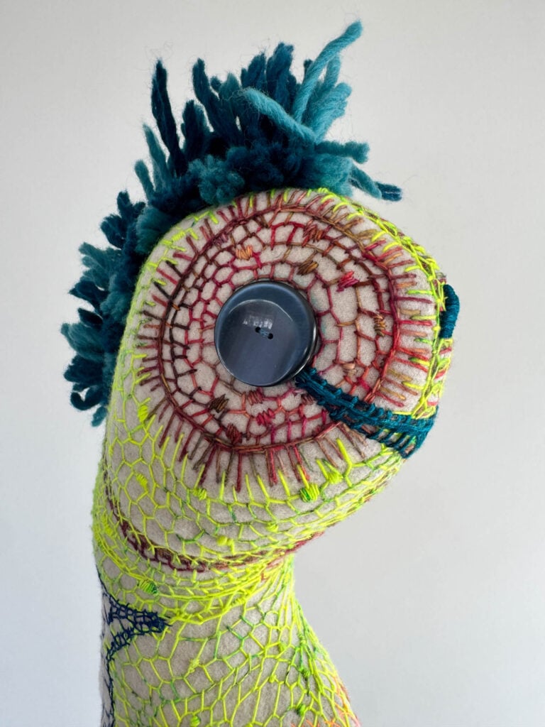 A soft sculpture by Wolfgang Woerner with neon yellow and red stitching, blue yarn tufts on top, and a large button-like eye on a textured, elongated head.