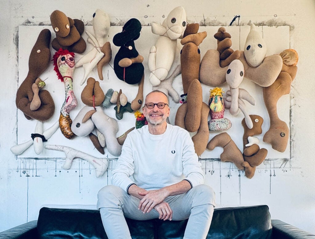 A man in light clothing sits on a dark couch, smiling, with a wall display of abstract, plush soft sculptures by Wolfgang Woerner behind him.