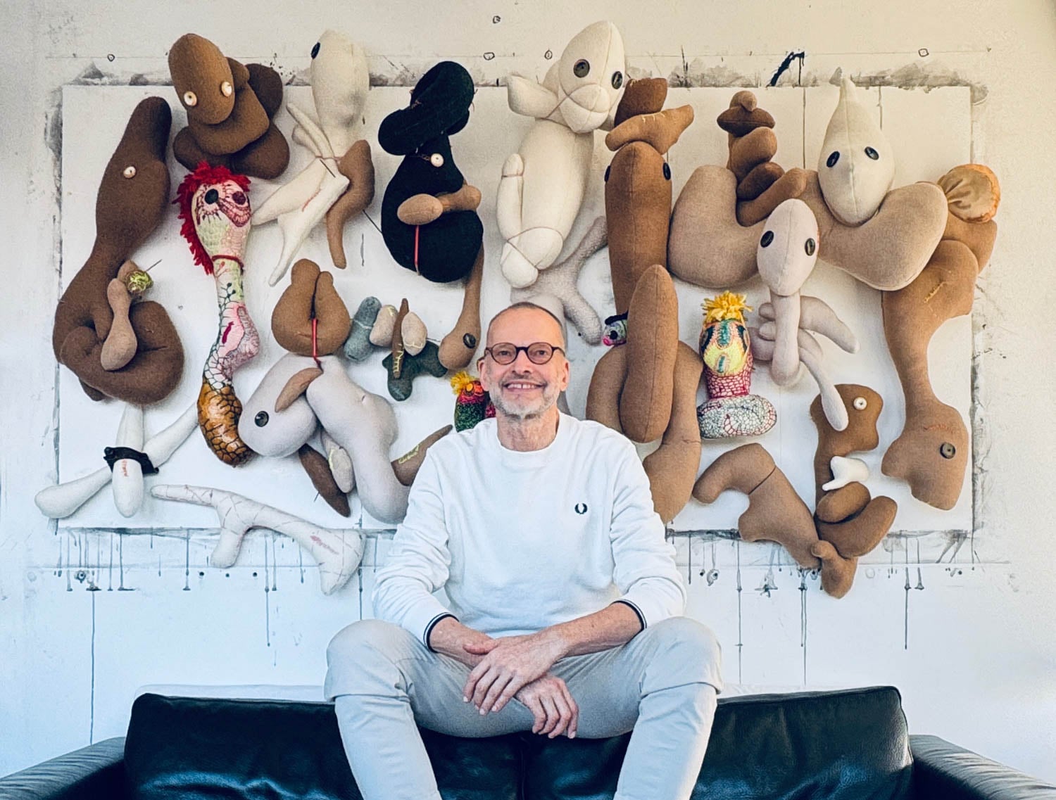 A man in light clothing sits on a dark couch, smiling, with a wall display of abstract, plush soft sculptures by Wolfgang Woerner behind him.