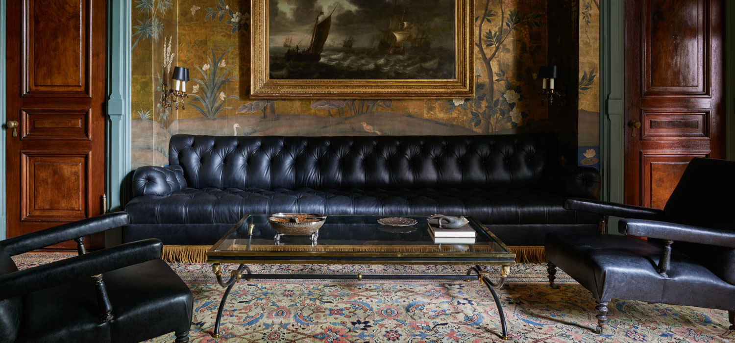 A room with a long black tufted leather sofa, glass coffee table, two black armchairs, and a patterned rug exudes timeless style—reminiscent of Billy Cotton’s signature touch—plus a large framed painting on a decorative wall.