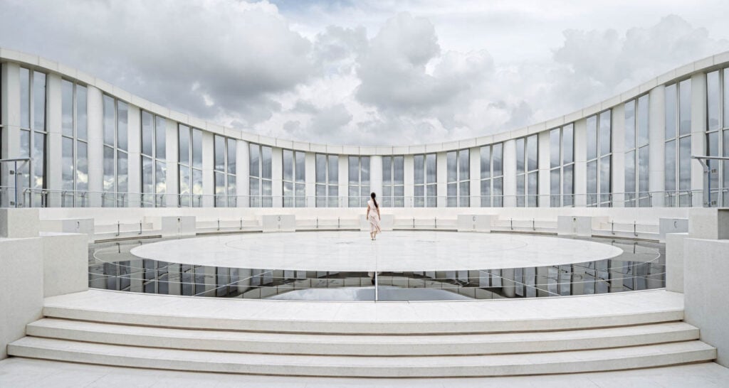 A lone person stands in the center of a circular, white, open-air structure with tall glass panels and cloudy sky in the background.