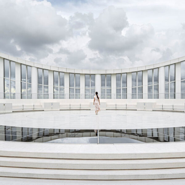 A lone person stands in the center of a circular, white, open-air structure with tall glass panels and cloudy sky in the background.