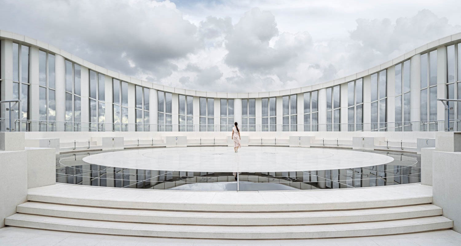 A lone person stands in the center of a circular, white, open-air structure with tall glass panels and cloudy sky in the background.