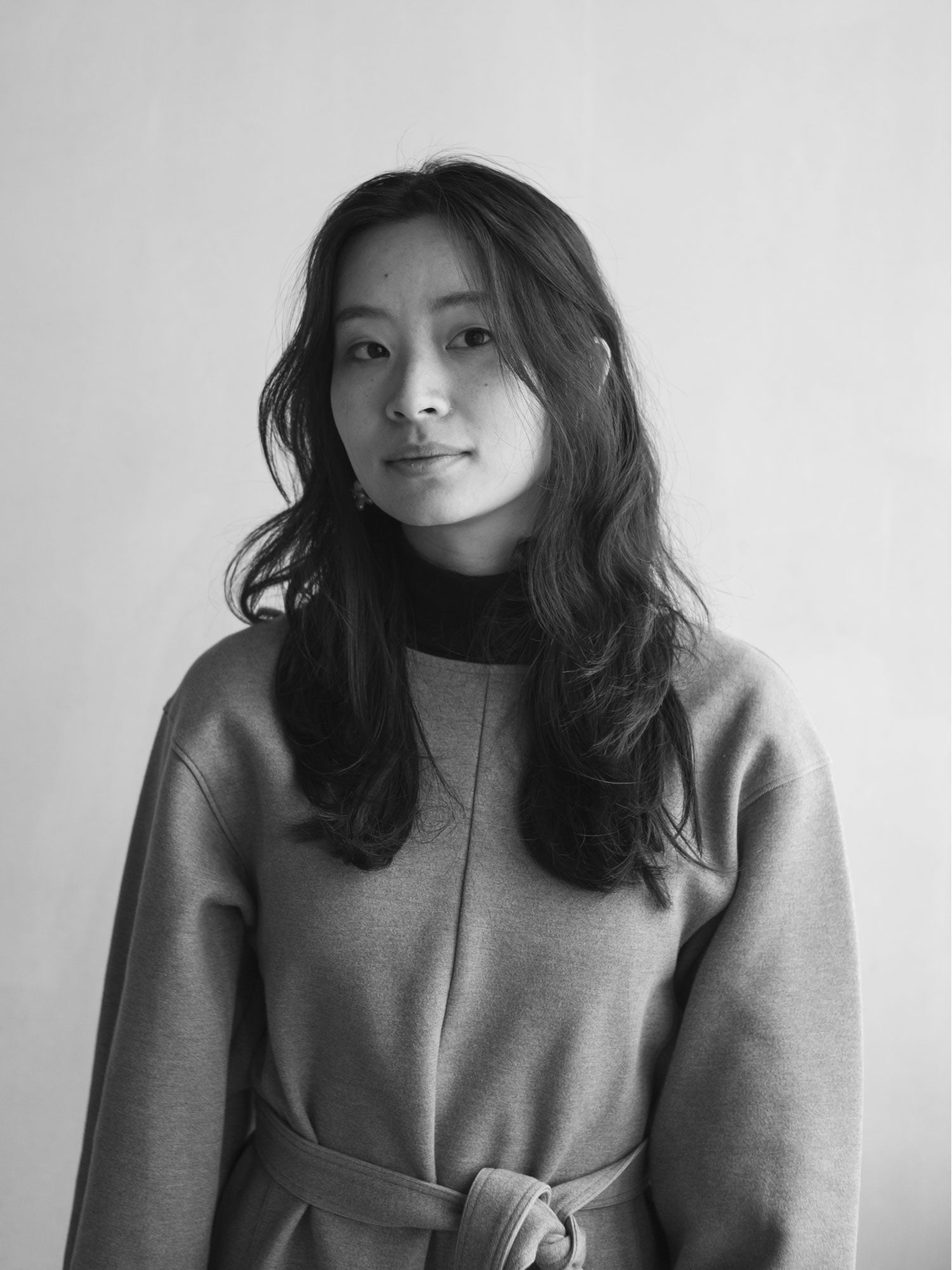 Black and white portrait of a woman with long hair, wearing a belted, long-sleeve top, standing in front of a plain background.