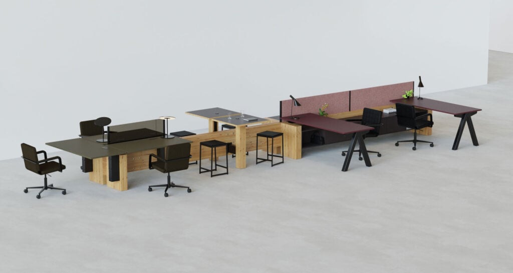 A row of modern office desks and chairs with partitions, desk lamps, and stools, arranged against a plain white wall on a light-colored floor.