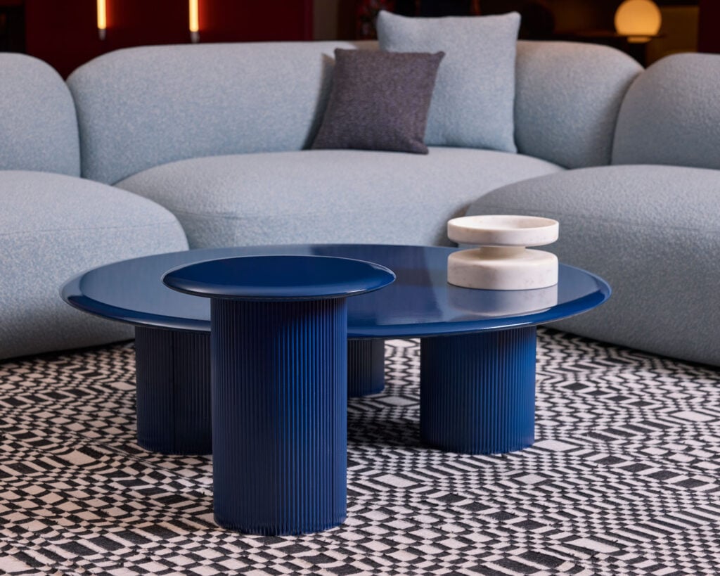 A modern living room with a light blue sofa, a round blue coffee table with ribbed legs, and two white marble decor pieces on a black and white patterned rug.