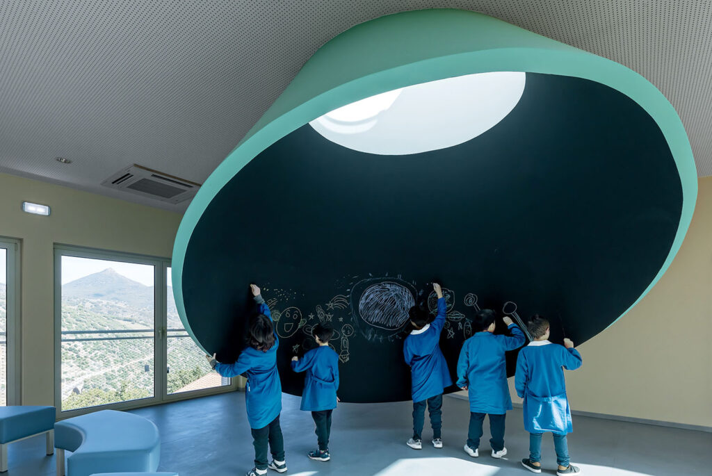 Five children in blue uniforms draw with chalk on a large, curved blackboard wall beneath a circular skylight in a bright room with mountain views.