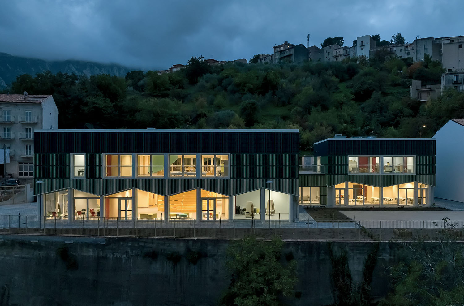 A modern two-story building with large illuminated windows stands against a hillside and cloudy evening sky, with trees and residential houses in the background.