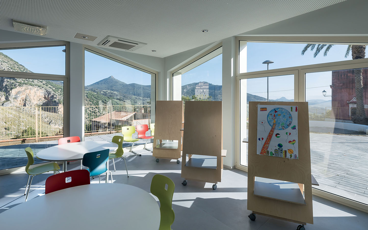 A bright classroom with round tables, colorful chairs, and large windows showing a mountain view. Two easels with artwork are positioned near the windows.