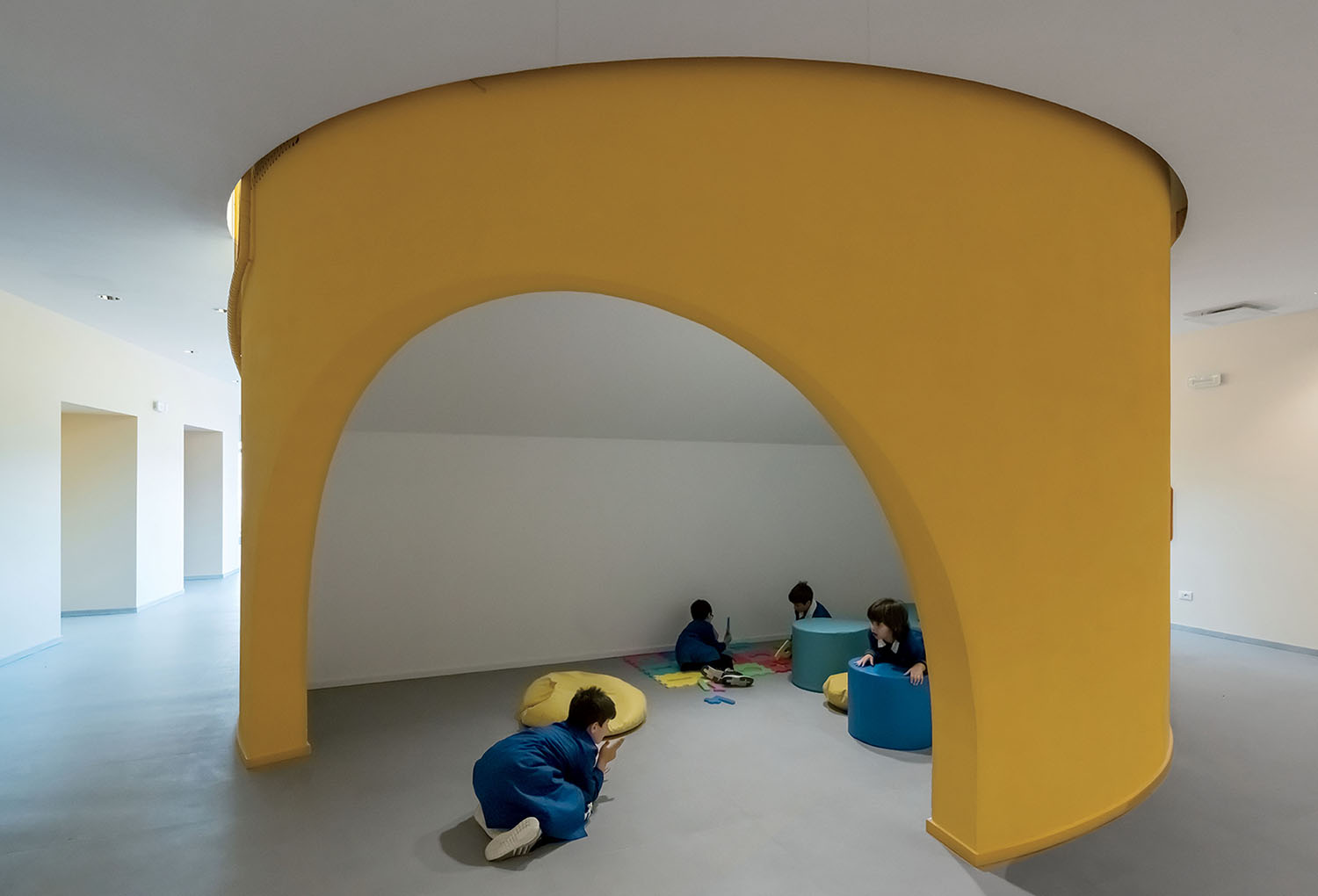 Children sit and play on the floor beneath a large yellow arch structure in a minimalist, modern indoor space with gray flooring and white walls.