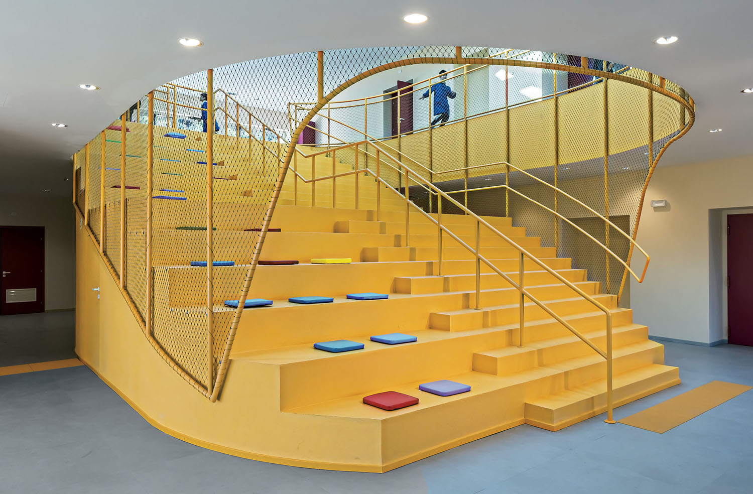 A modern yellow staircase with integrated seating steps and colorful cushions, surrounded by a yellow mesh railing, in a brightly lit indoor space.