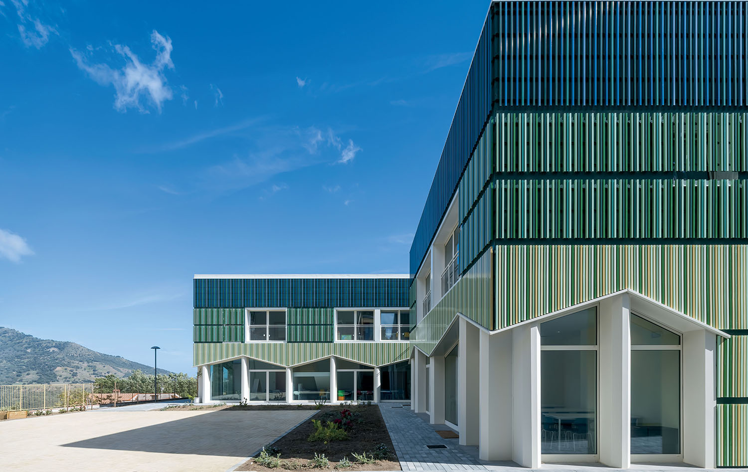 Modern two-story building with green and white vertical panels, large windows, and a geometric roofline under a clear blue sky. A mountain is visible in the background.