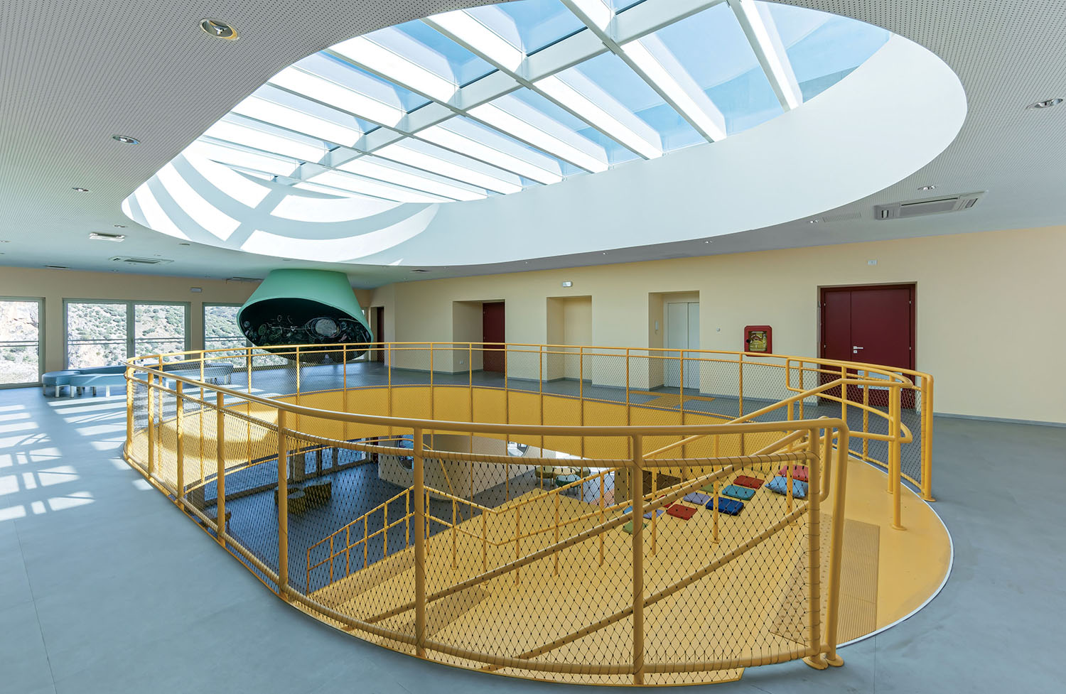 Interior of a modern building with a yellow spiral staircase, red doors, large windows, and an oval skylight letting in natural light.