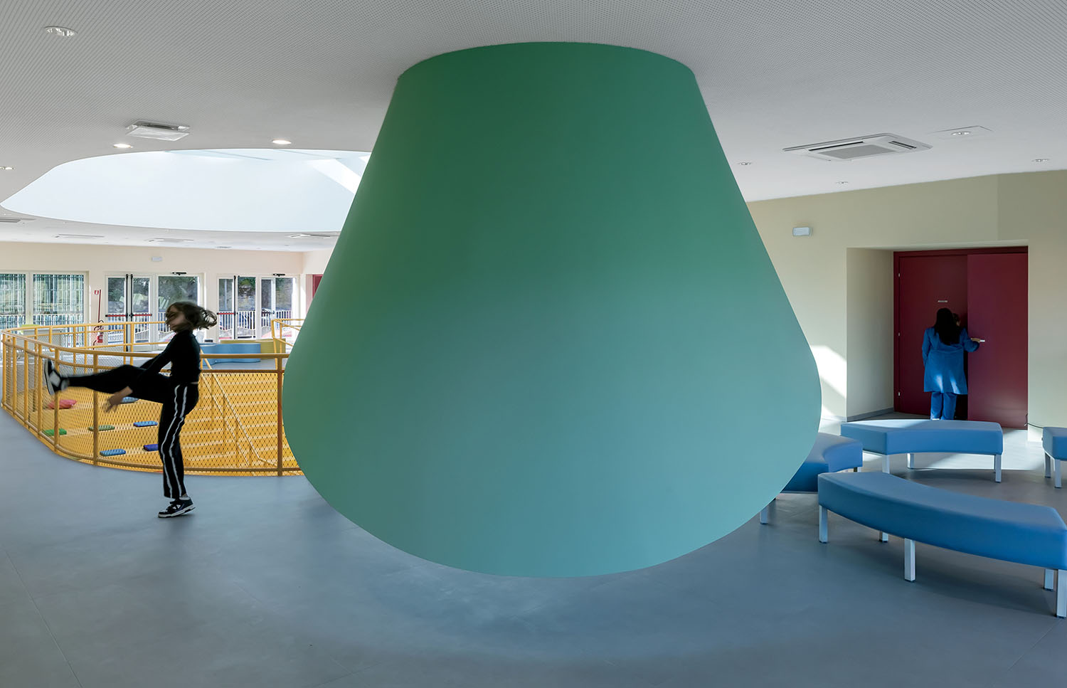 A large green cone hangs from the ceiling in a modern, open indoor space with curved benches; one person is kicking a ball, another stands near a red door.
