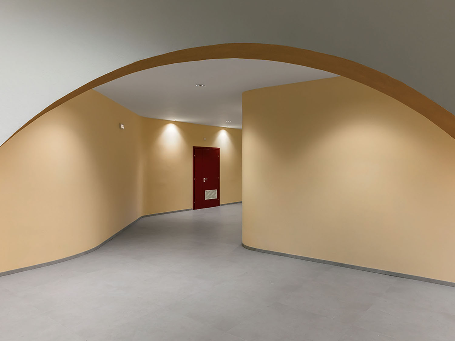 A modern hallway with beige curved walls, a grey floor, and a red door at the end, illuminated by recessed ceiling lights.