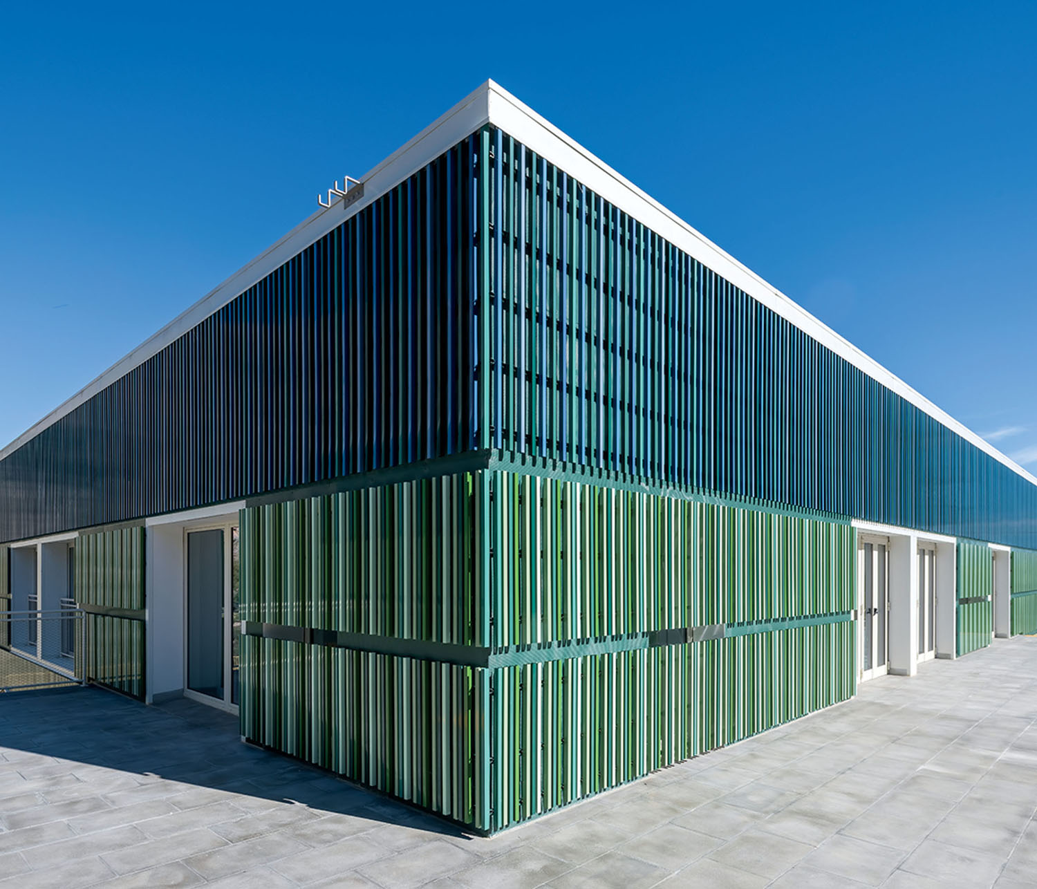 A modern building with green and blue vertical slats on its exterior, flat roof, and a paved area in the foreground under a clear blue sky.