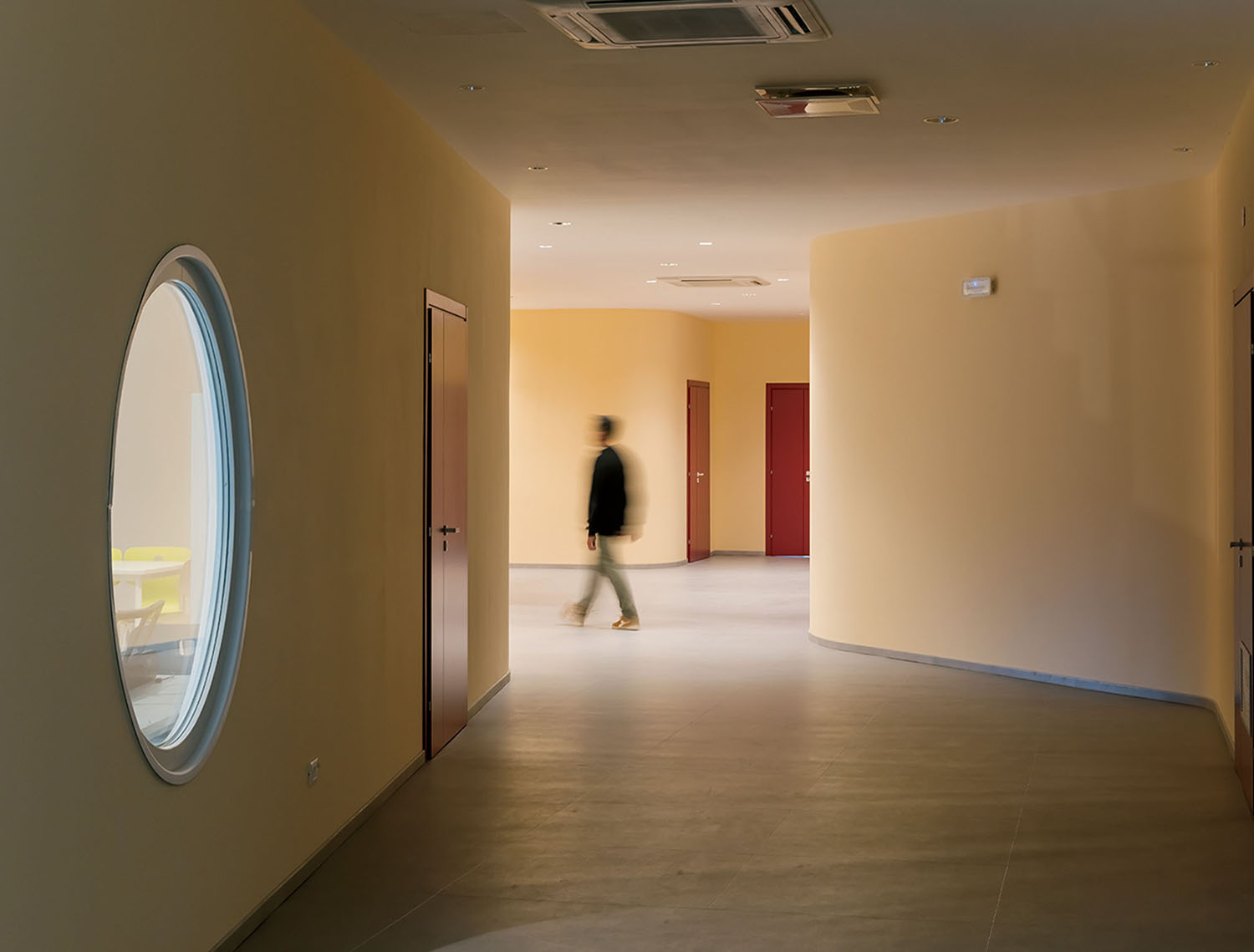 A person walks through a hallway with beige walls, red doors, and a circular window on the left side.