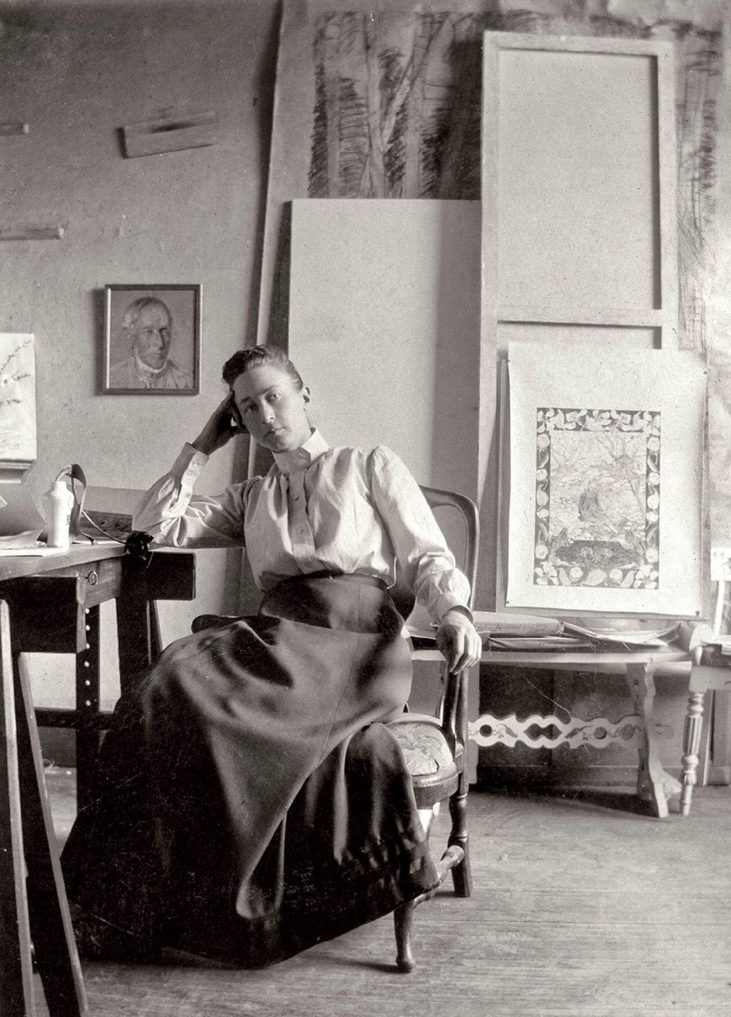 A woman in early 20th-century attire sits in an art studio, resting her head on her hand, surrounded by framed artworks, sketches, and artistic materials.