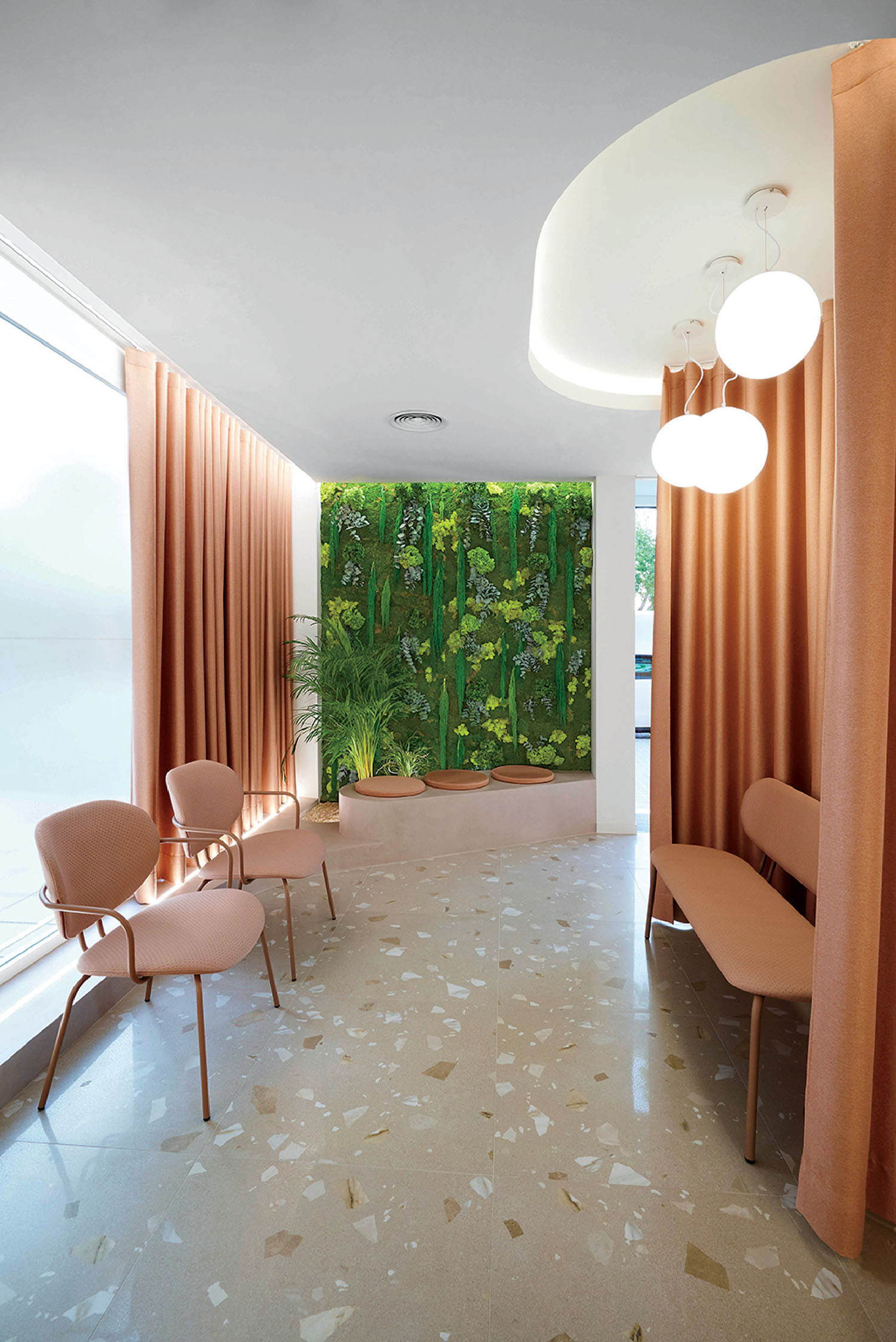 A modern waiting room with pink chairs, a terrazzo floor, peach curtains, round ceiling lights, and a green vertical garden on the back wall.