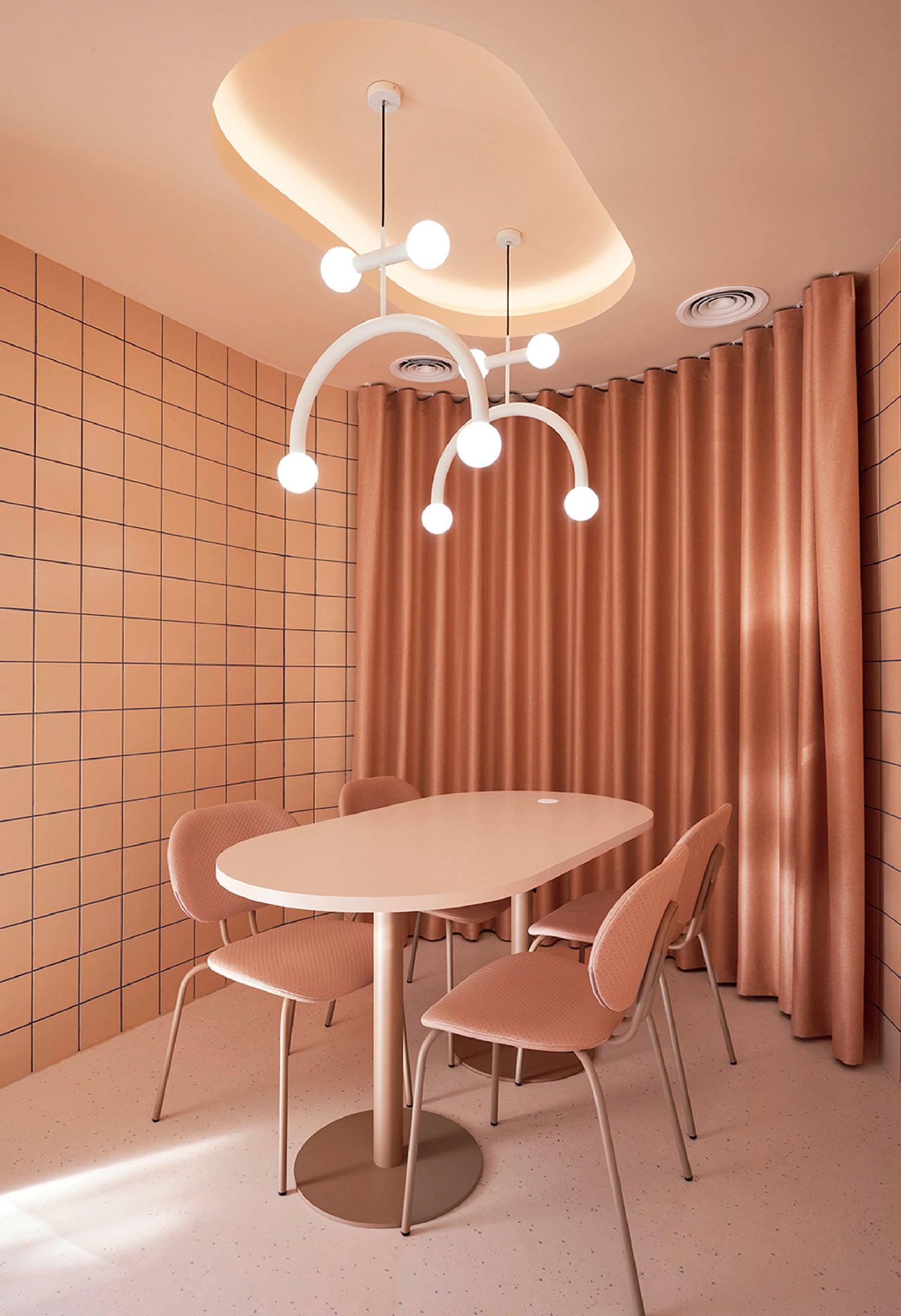 A minimalist meeting room with a rounded table, four pink chairs, peach-colored tiled walls, and a modern chandelier, with peach curtains covering one wall.