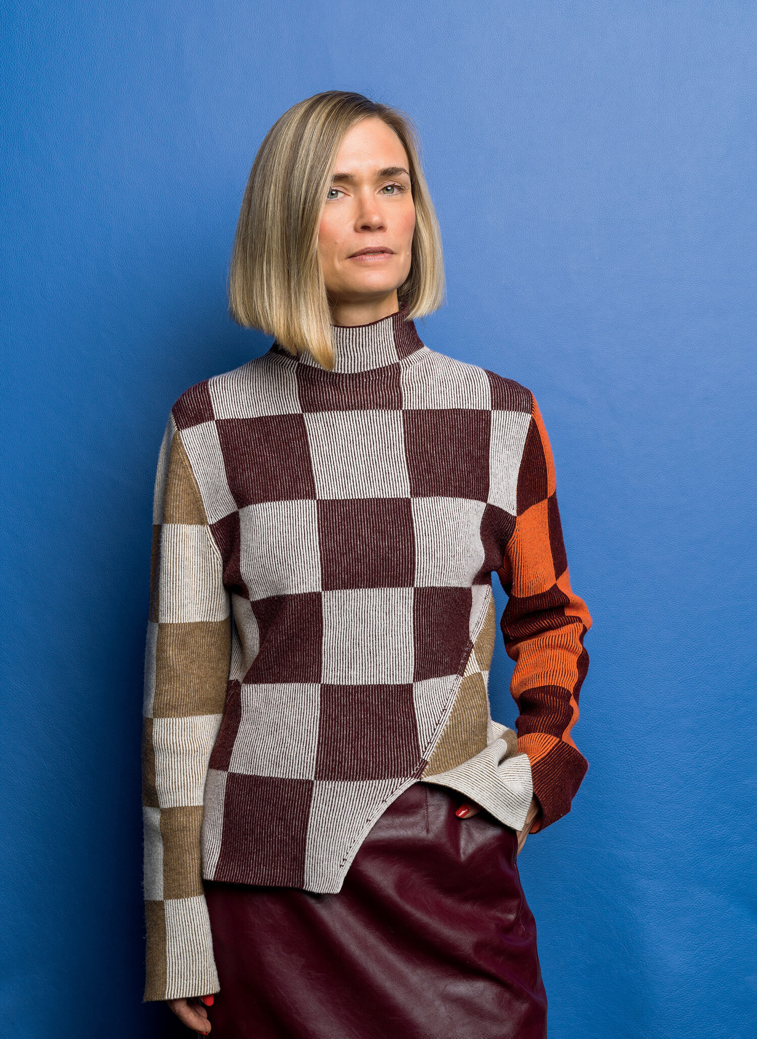 A person with straight, shoulder-length blond hair stands against a blue background, wearing a checkered turtleneck sweater and a burgundy skirt, with one hand in their pocket.