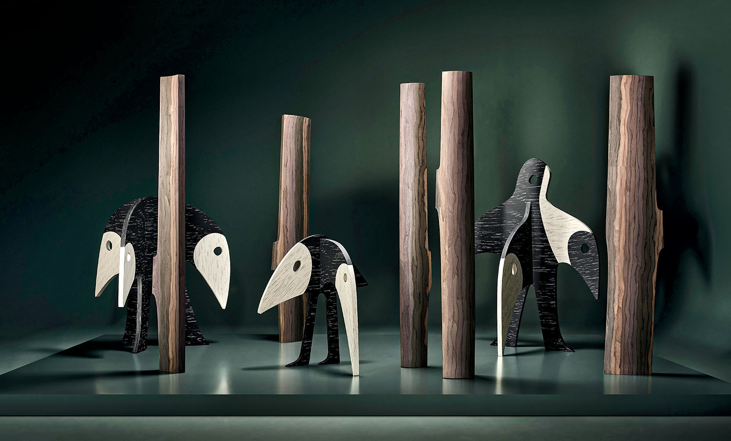 Several abstract, bird-like sculptures with black and white surfaces stand among vertical wooden columns on a green background.