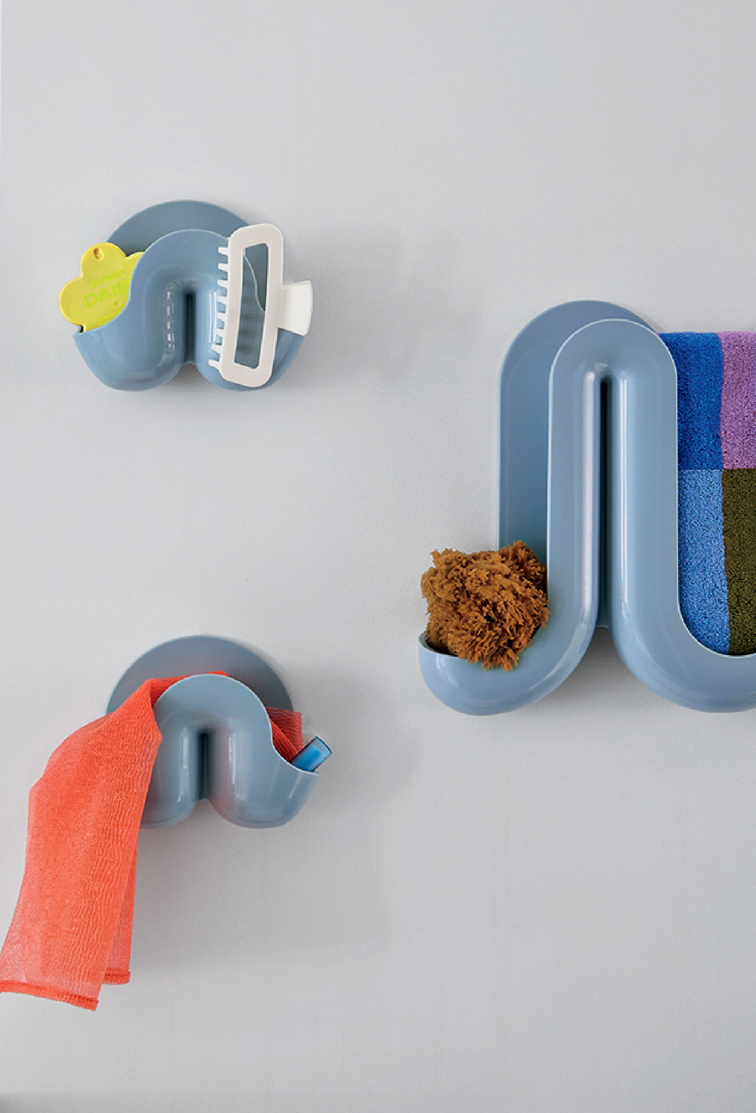 Three blue, wall-mounted bathroom holders display items: a sponge, a towel, a scrub brush, and a colorful washcloth, all against a light grey background.
