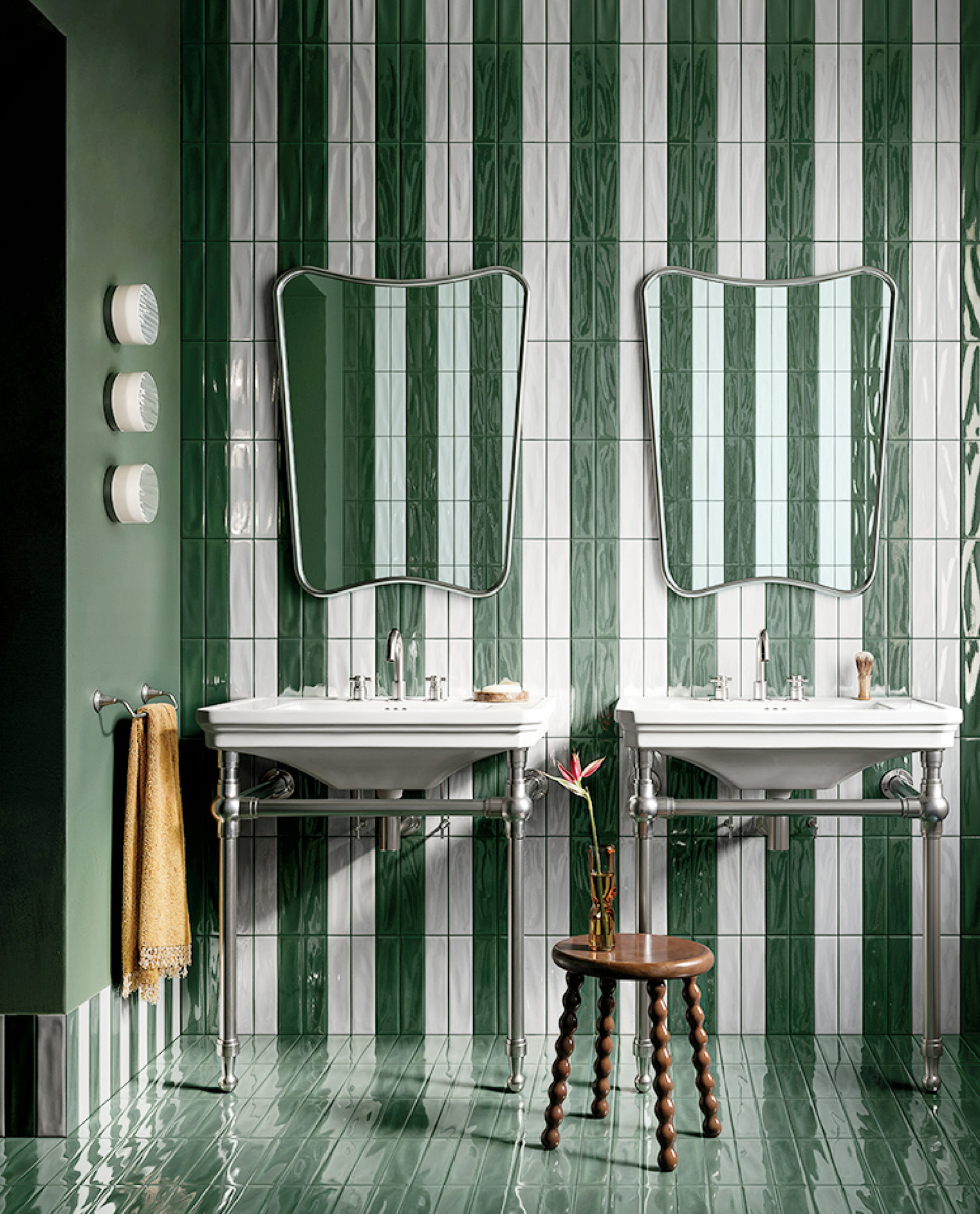 A bathroom with green and white vertical striped tiles, two sinks with mirrors, a small wooden stool, and a towel hanging on the left wall.