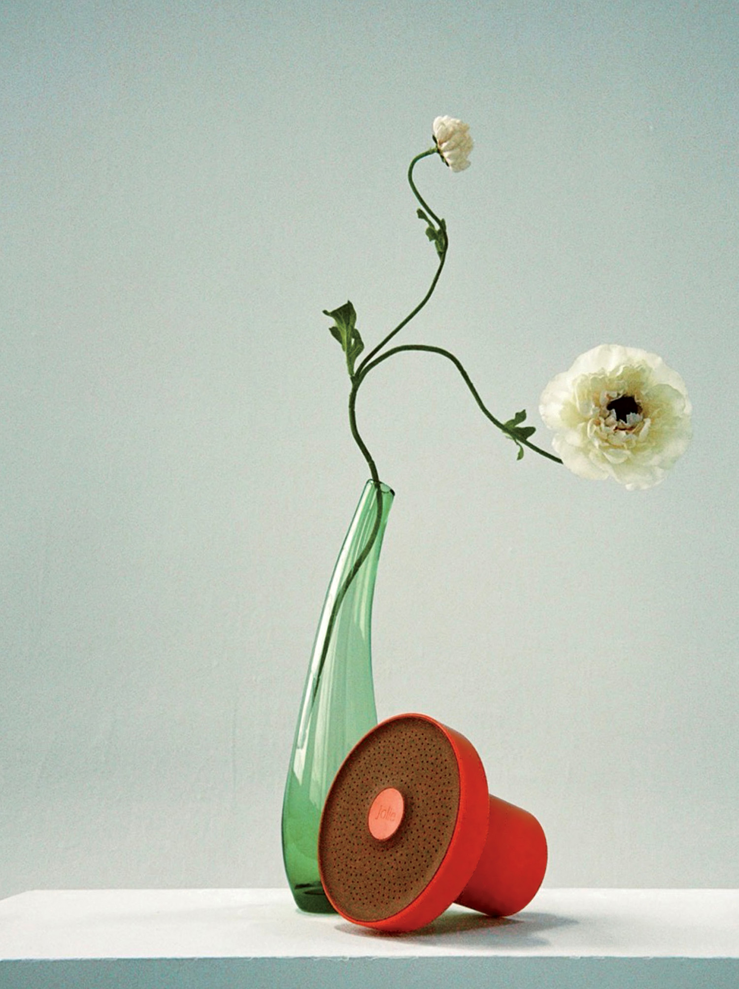 A green glass vase with two white flowers and a red portable speaker are arranged on a white surface against a plain light gray background.