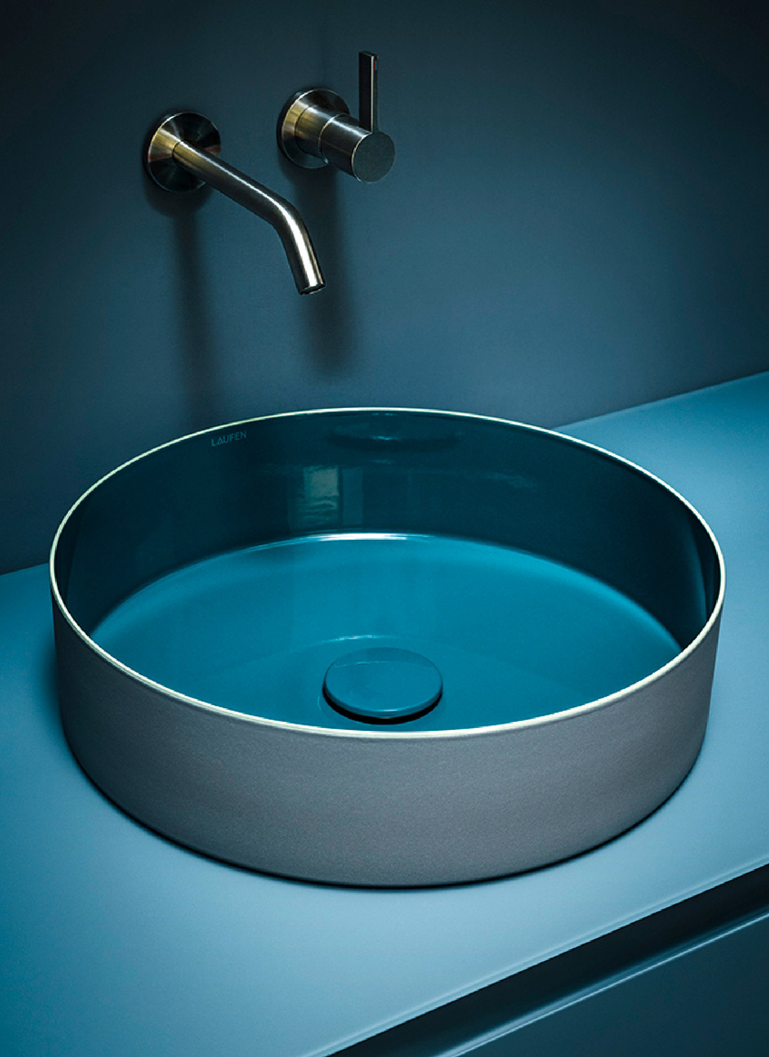A modern, round vessel sink with a blue interior and gray exterior, paired with a wall-mounted metal faucet, set against a matching blue countertop and wall.