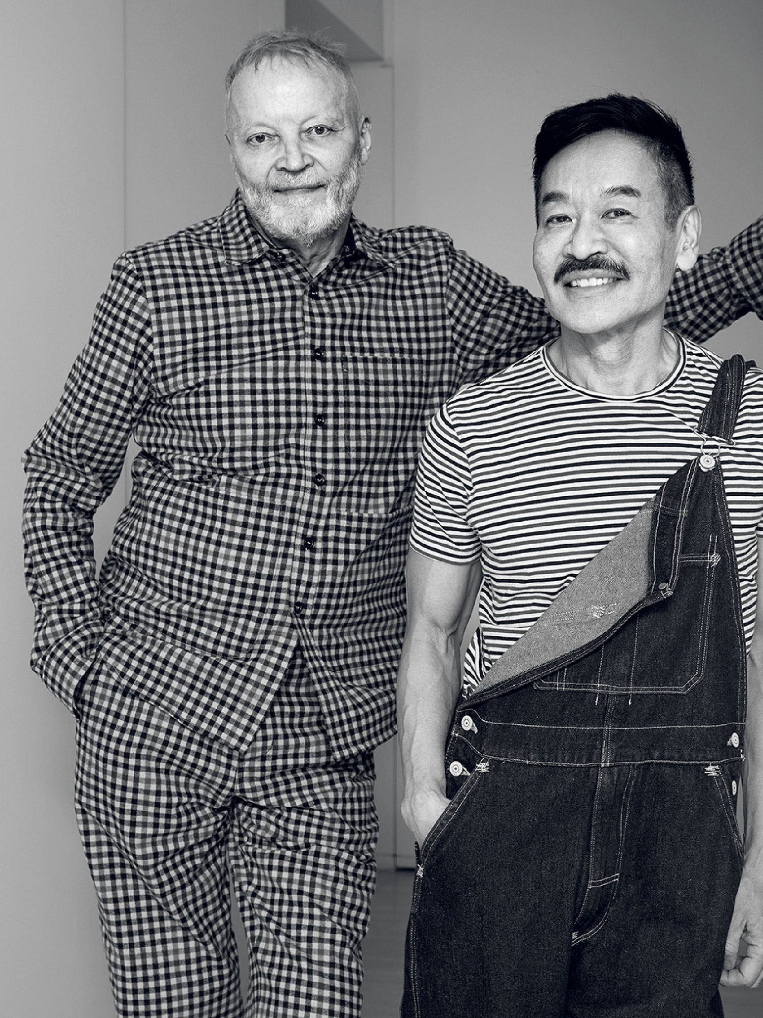 Two men pose together indoors; one wears a checkered outfit and the other wears a striped shirt with denim overalls.