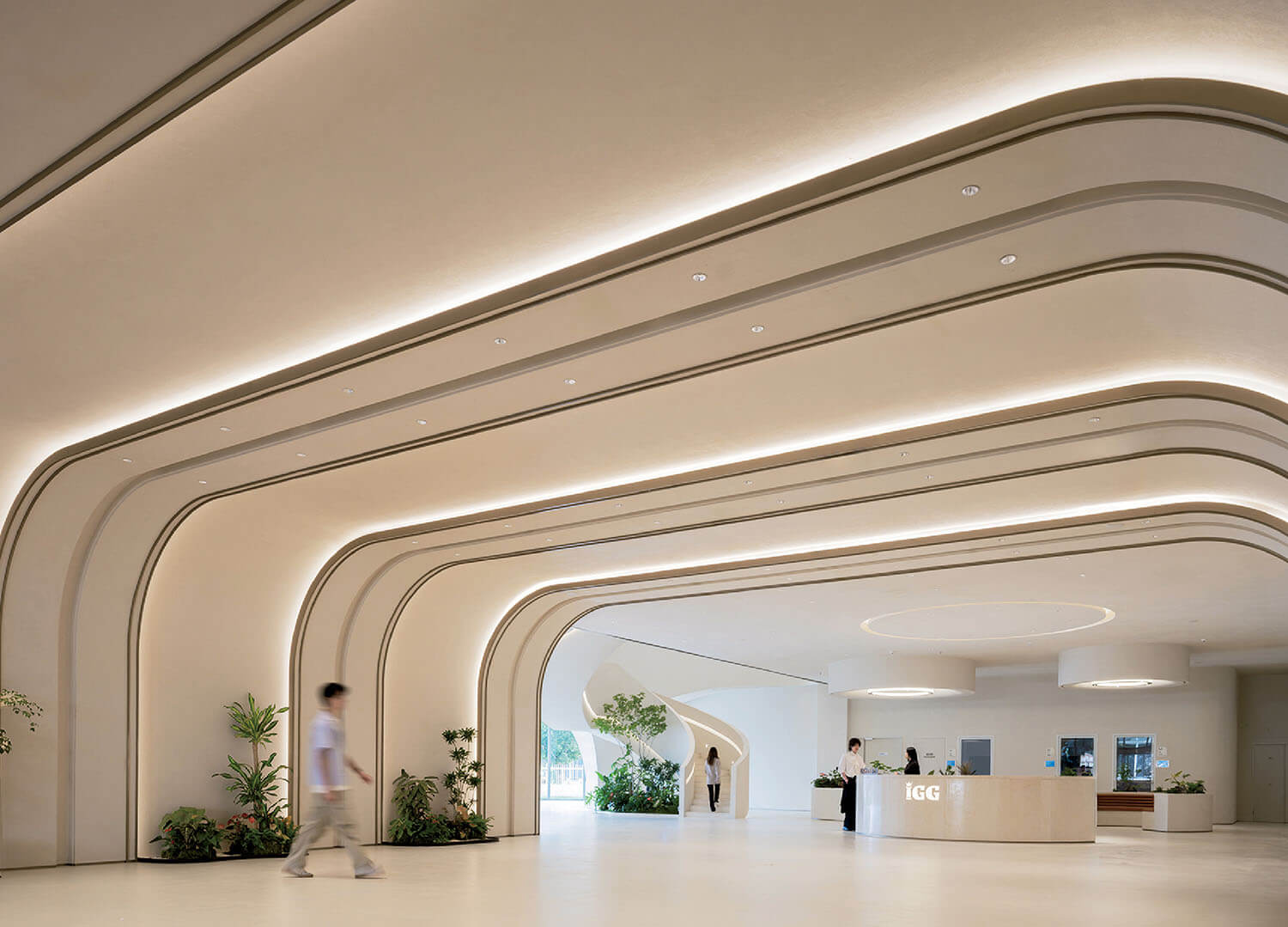 Spacious, modern lobby with curved ceiling panels, recessed lighting, indoor plants, and a reception desk; a person walks across the floor.