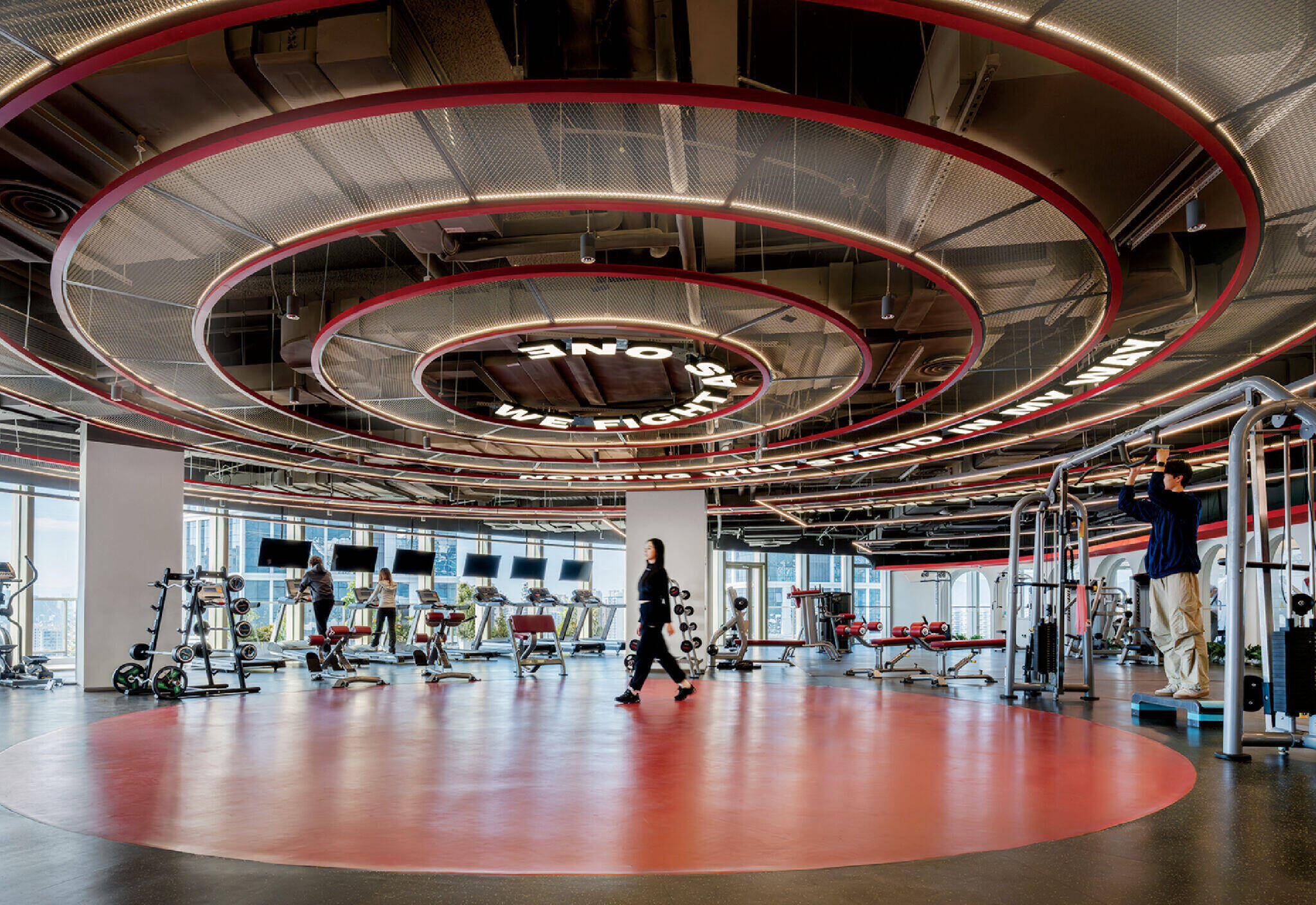 Modern gym interior with circular ceiling lights, exercise machines, free weights, and people working out on various equipment.