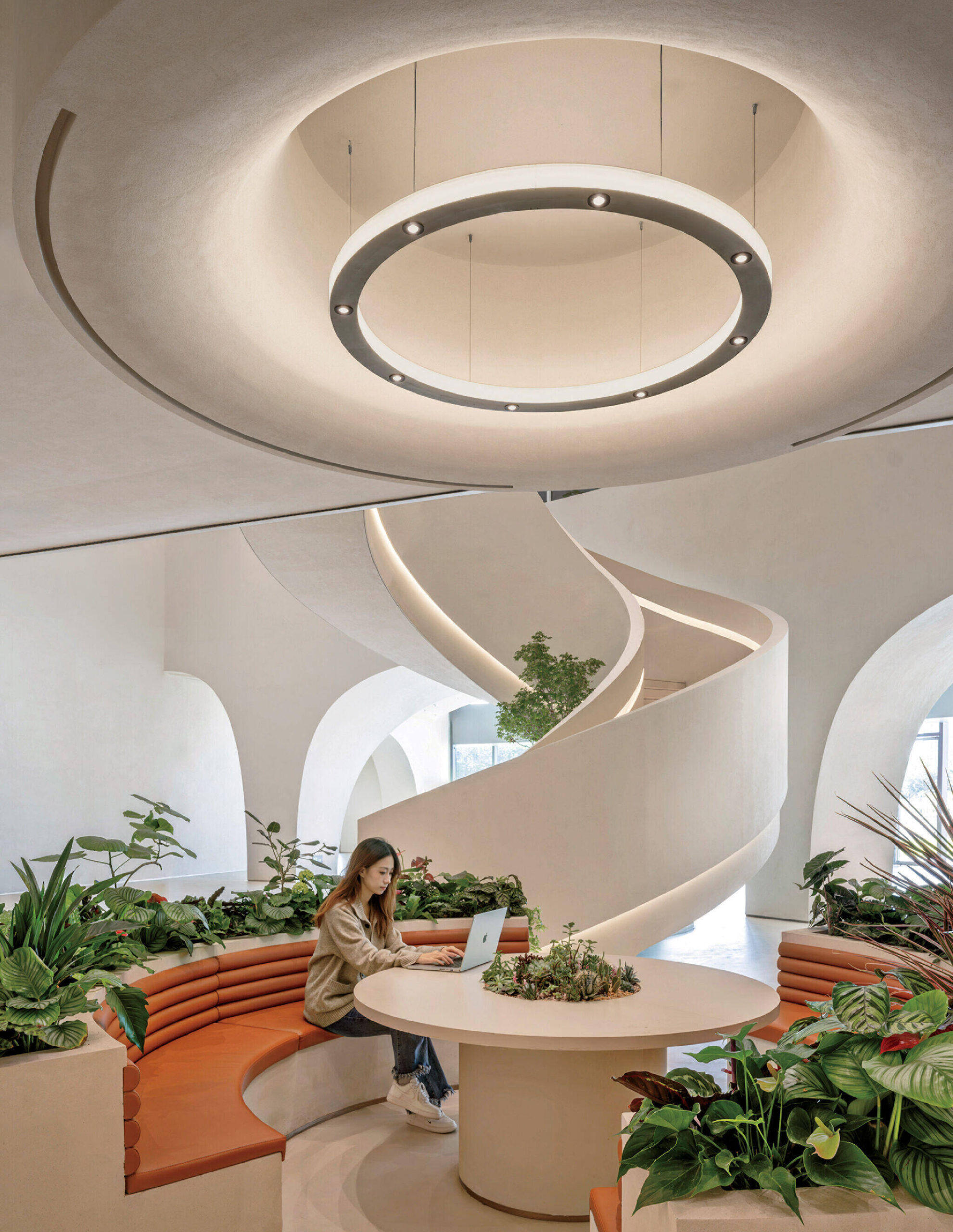 A woman works on a laptop at a round table in a modern, white interior with a spiral staircase, circular light fixture, curved seating, and surrounding plants.