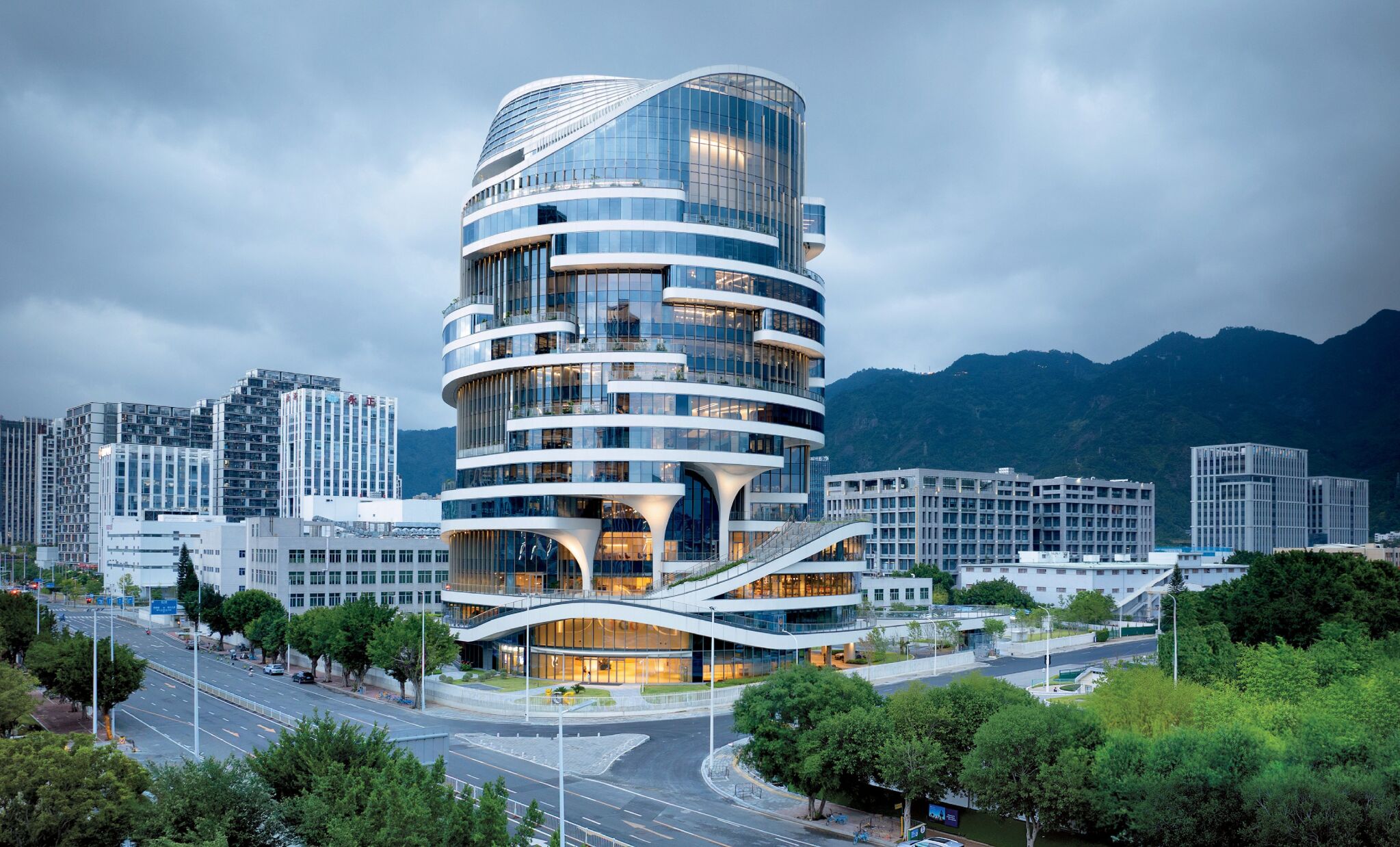 A modern, multi-story building with curved, white architectural features stands among other buildings, with mountains visible in the background.
