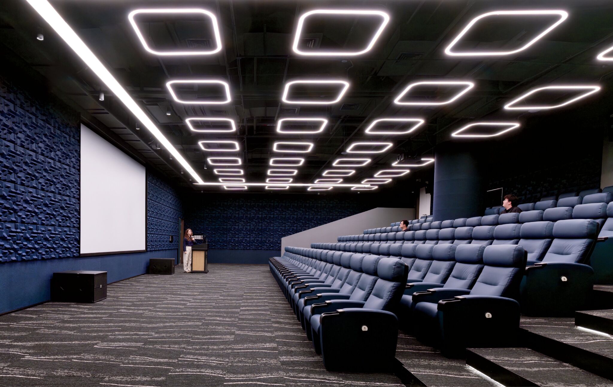 A modern lecture hall with blue cushioned seats, geometric ceiling lights, a large projector screen, and a speaker standing at a podium. Few people are seated.