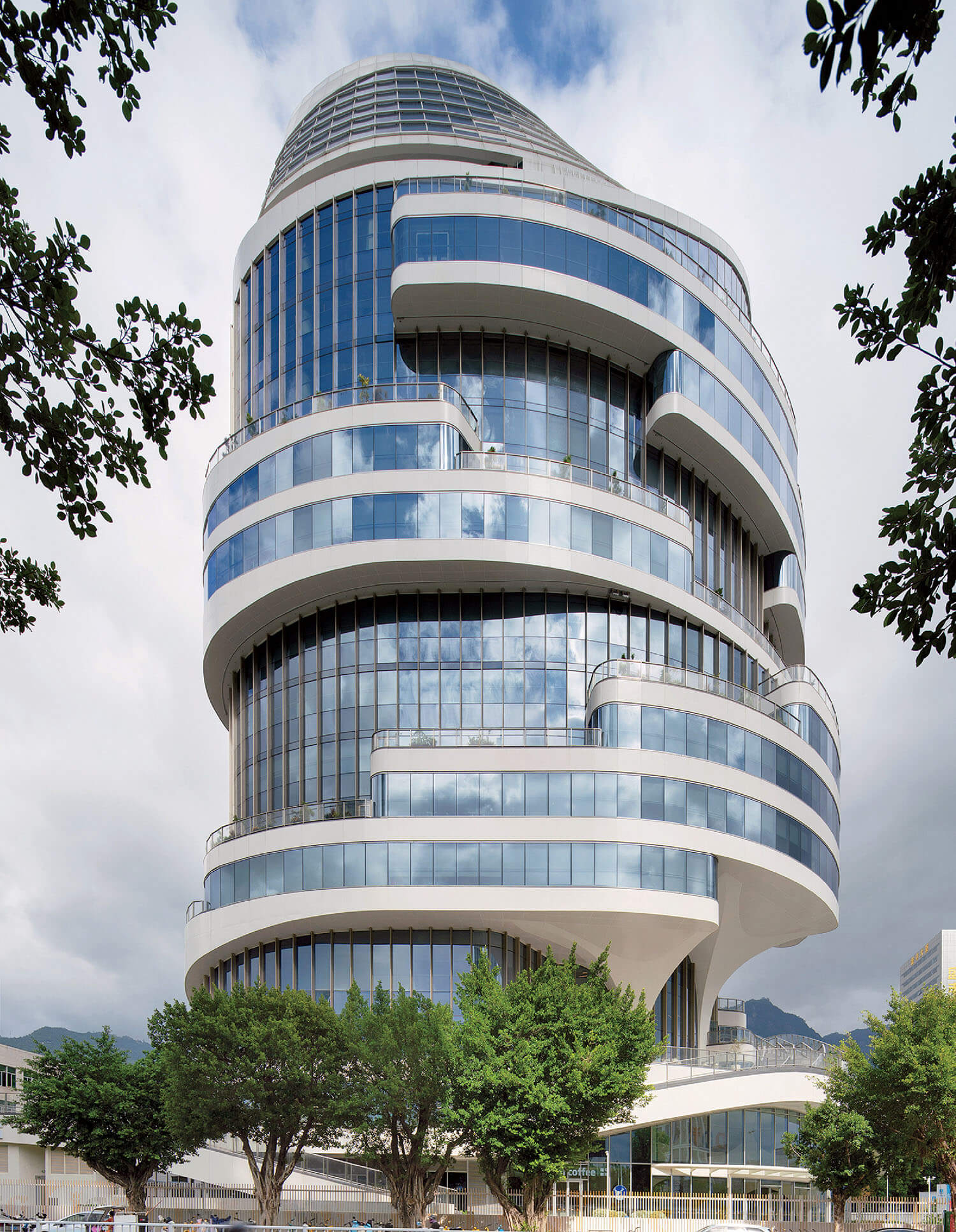 Modern multi-story building with curved, layered glass and white exterior, featuring prominent horizontal bands and surrounded by trees.