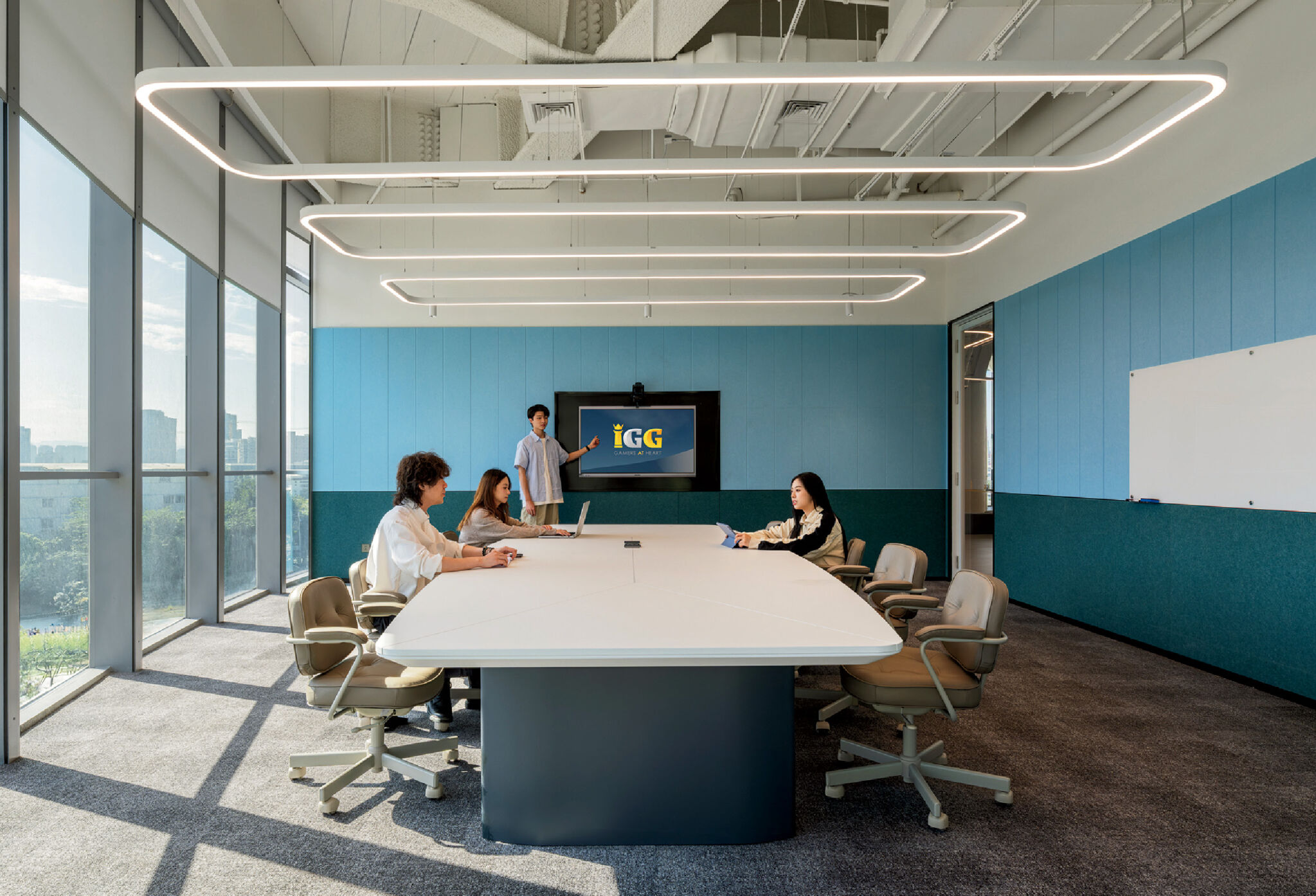 Four people sit around a large conference table in a modern meeting room with floor-to-ceiling windows; one person stands near a screen displaying a presentation.