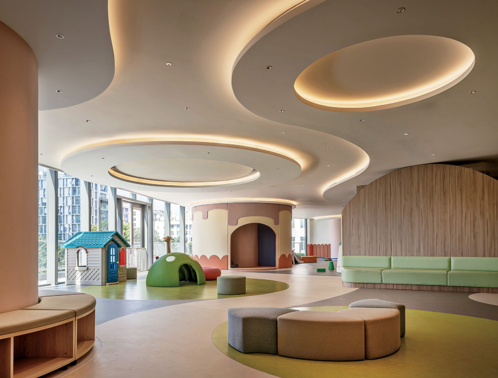 Spacious, modern indoor playroom with curved ceiling lights, soft seating, a toy house, and large windows letting in natural light.