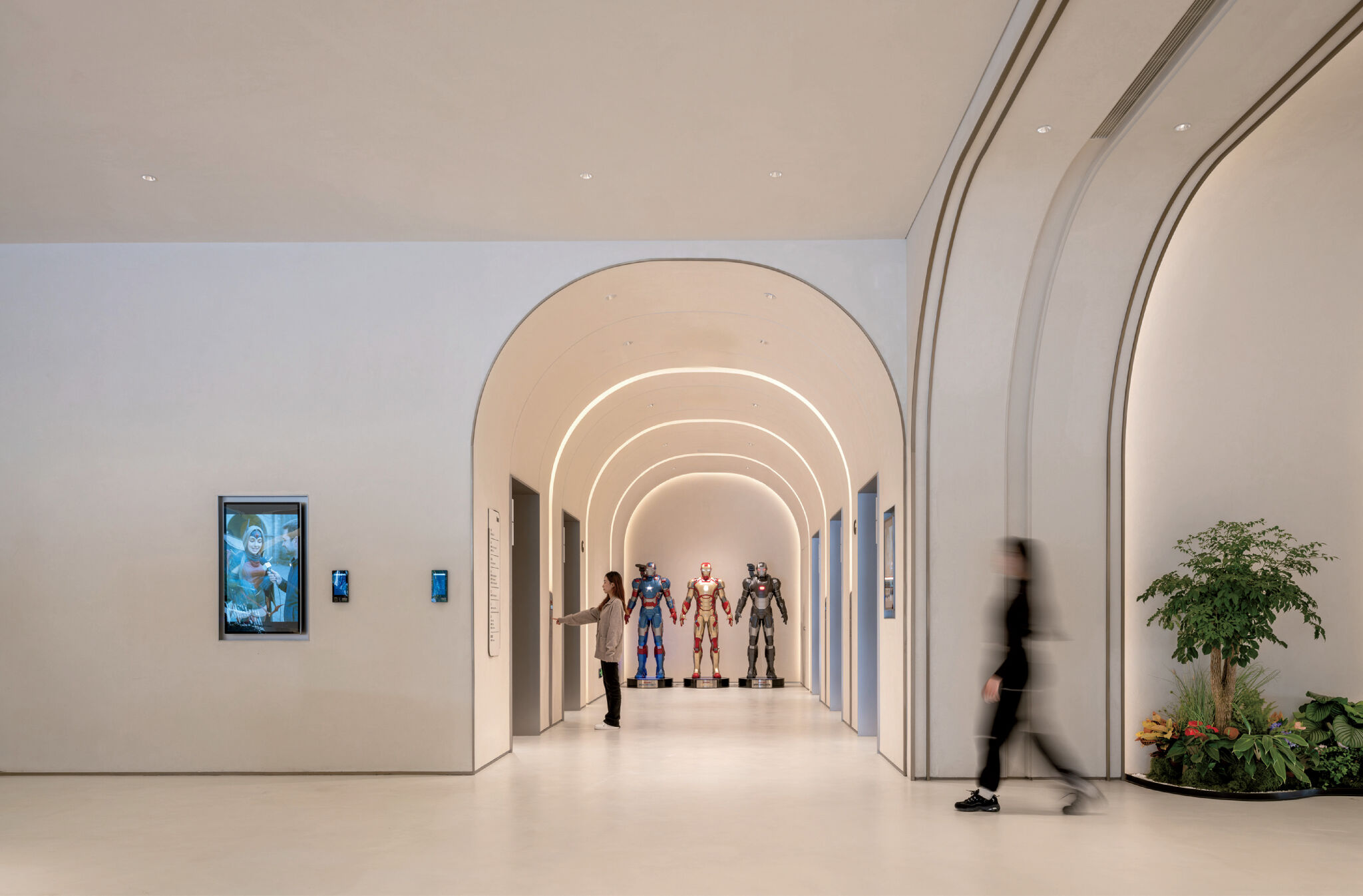 A modern hallway with arched ceilings features a person gesturing toward three superhero statues, while another person walks past a plant and digital screen.