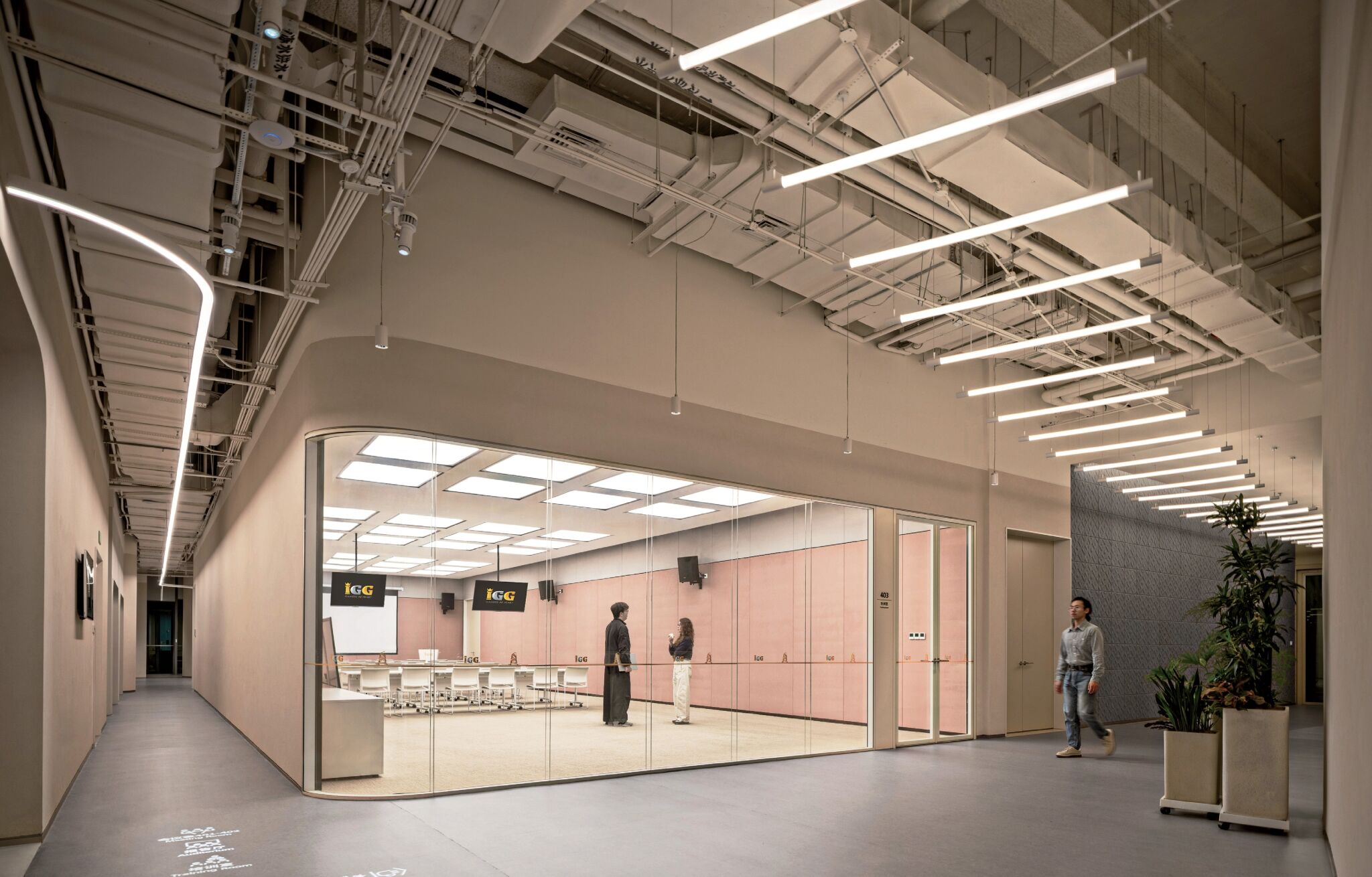 A modern hallway with exposed ceiling lights and pipes, adjacent to a glass-walled conference room with people inside and a person walking past outside.