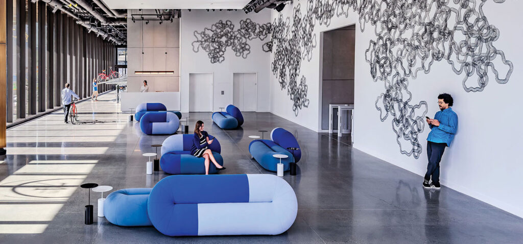 Spacious modern lobby with blue seating, abstract wall art, large windows, and people sitting, walking, and standing.
