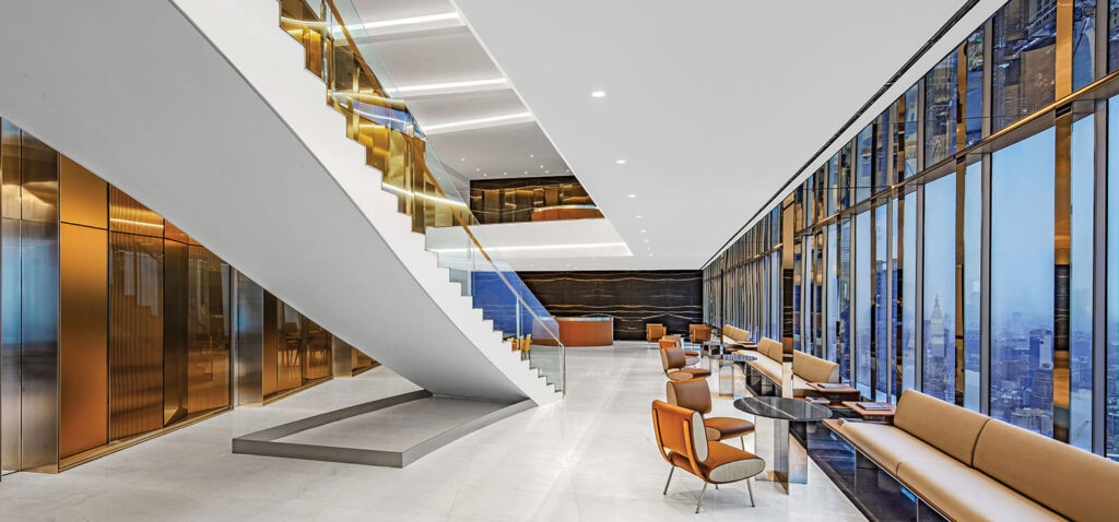 Modern office lounge with floor-to-ceiling windows, tan seating, black tables, and a white staircase—reflecting the sophisticated ambiance often found in Covington & Burling’s workspaces. City skyline is visible through the glass walls.