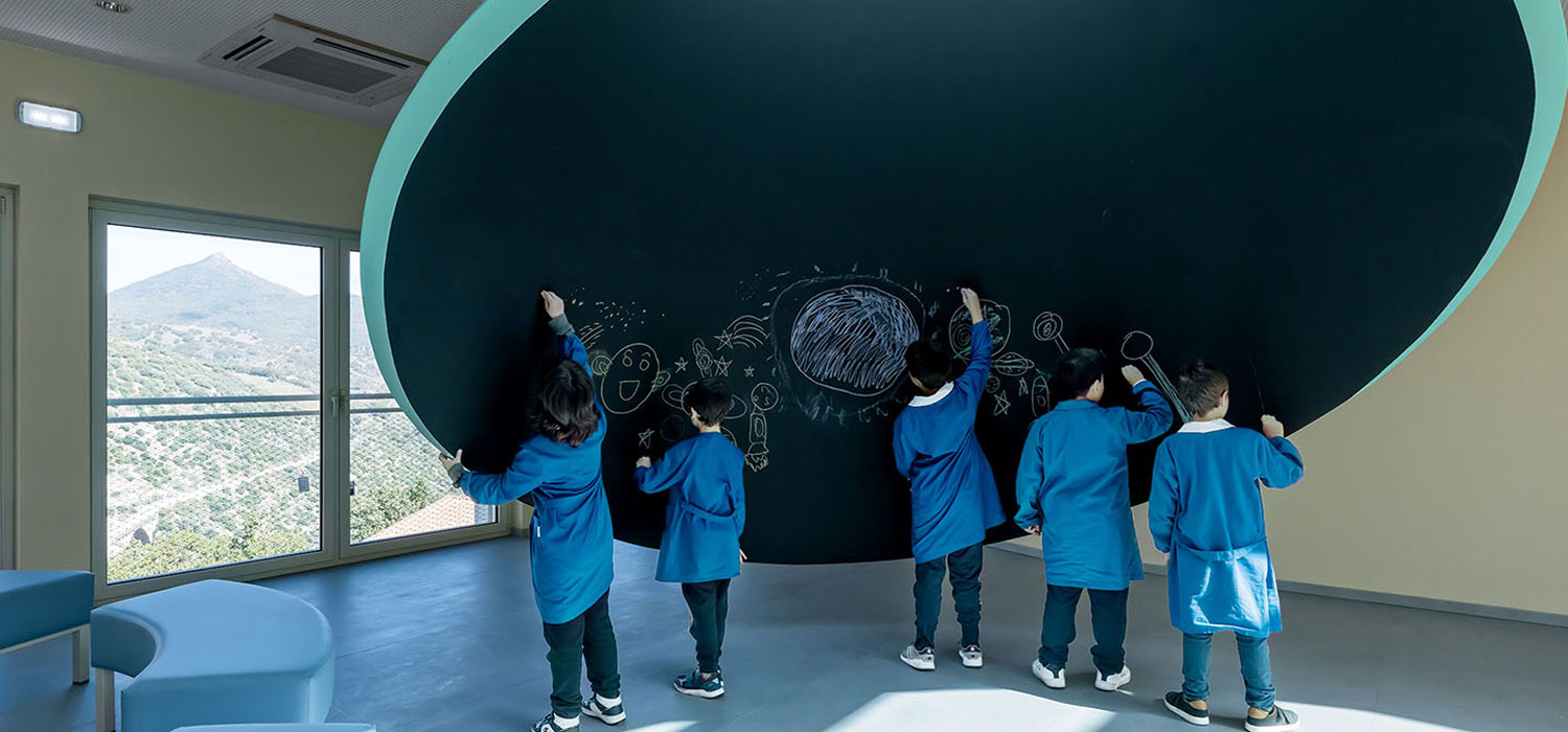 Five children in blue uniforms draw with chalk on a large, curved blackboard in a brightly lit classroom with a mountain view through the window.