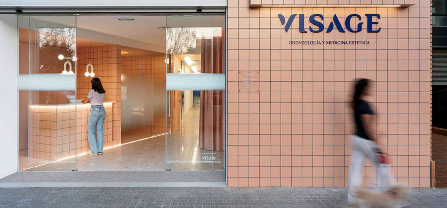 Modern clinic entrance with peach tile walls, glass doors, and the sign "VISAGE Odontología Y Medicina Estética." One person stands at the reception, another walks outside with a dog.