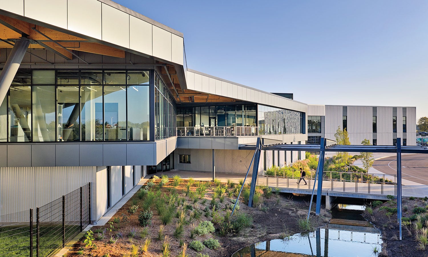 Modern two-story building with large windows, a covered walkway, landscaped grounds, and a reflective water feature in the foreground.