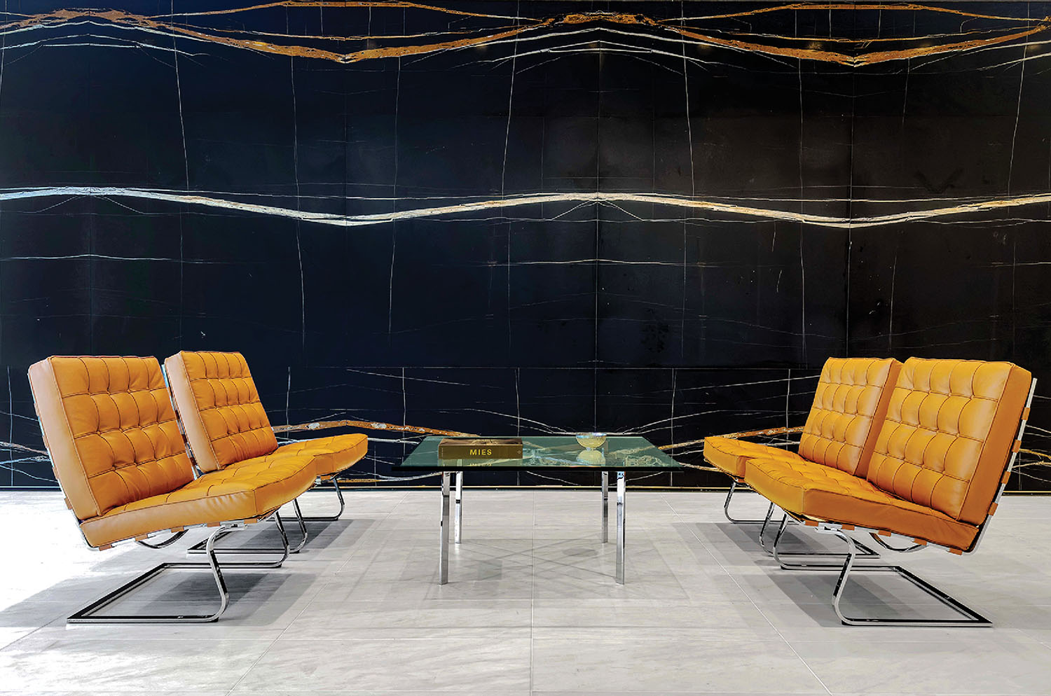 Four orange leather chairs with chrome legs face each other around a glass coffee table, set against a black marble wall with white and gold veins.